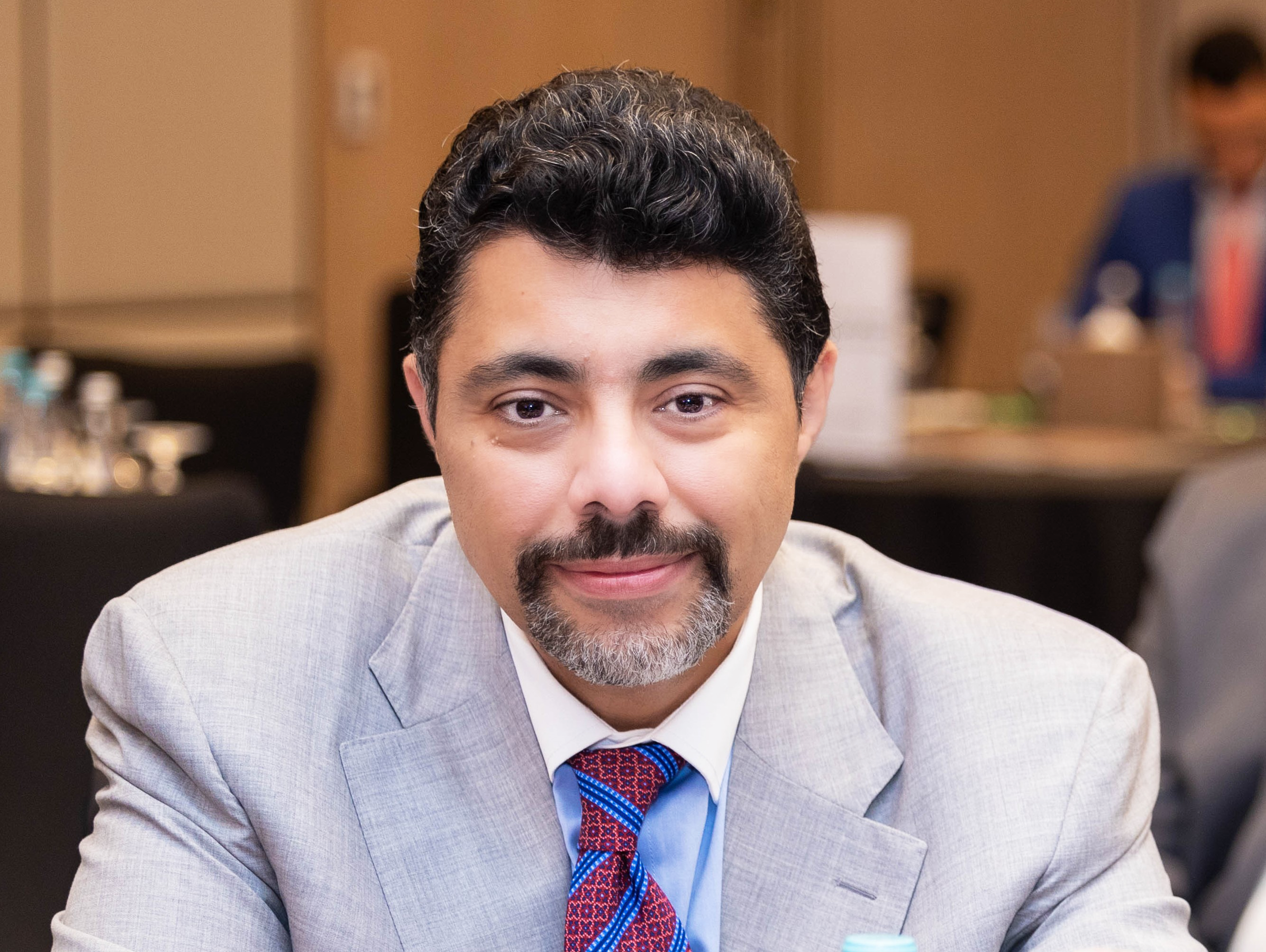 Man with dark hair wearing light grey suit and tie, smiling into camera